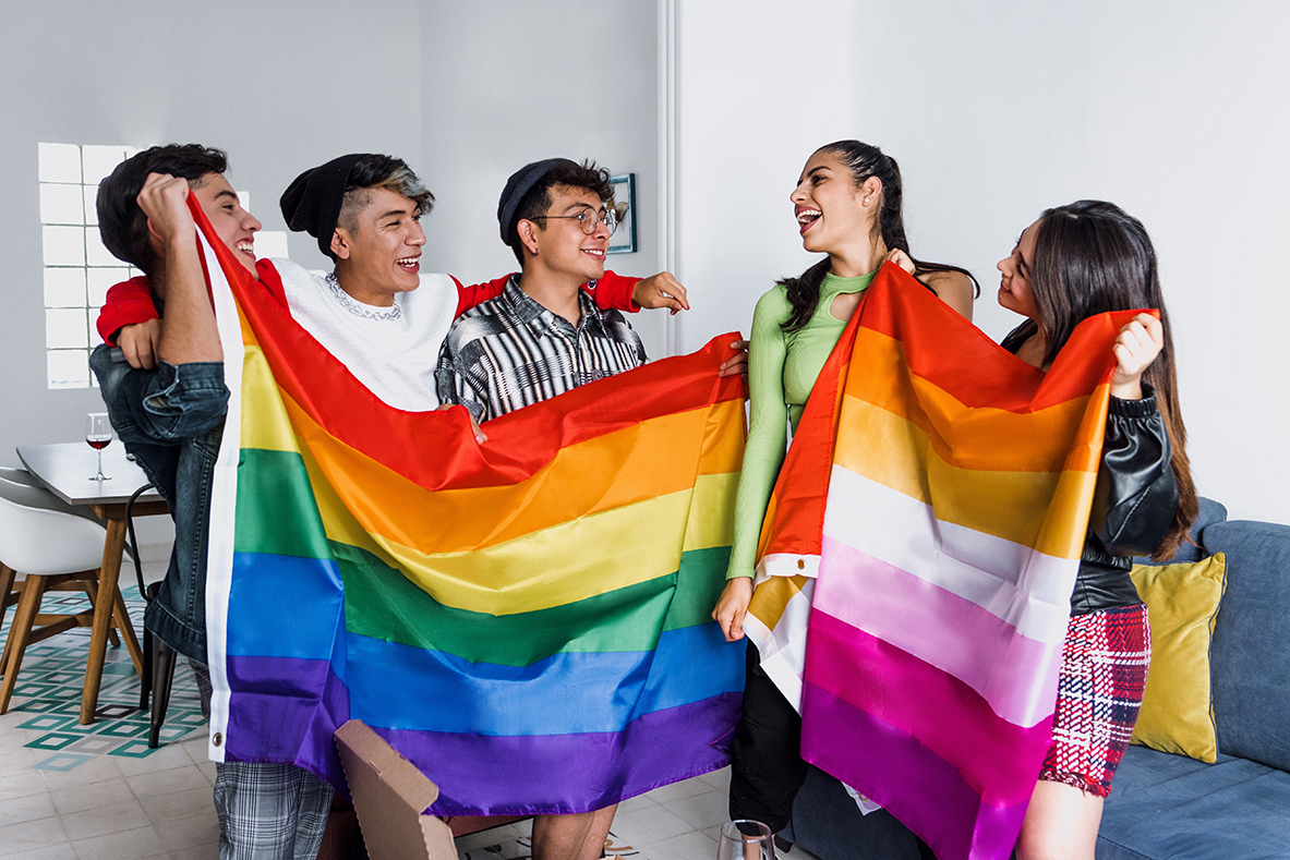 Young Latin LGBTQIA friends celebrating gay pride month holding rainbow flag at home in Mexico. GLFHC offers Gender-Affirming Care.