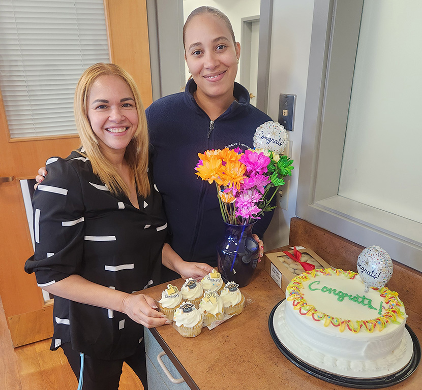 Two GLFHC Pharm Tech graduates from the Regis College training program pose with cake, cupcakes and flowers to celebrate their achievement.
